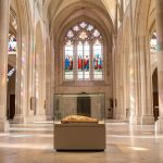 Comment explorer l’histoire et les controverses de la Tunique d’Argenteuil ? Vue intérieure lumineuse d’une basilique historique avec des vitraux colorés et une relique textile présentée sobrement sous verre protecteur.