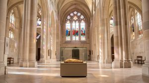 Vue intérieure lumineuse d’une basilique historique avec des vitraux colorés et une relique textile présentée sobrement sous verre protecteur.