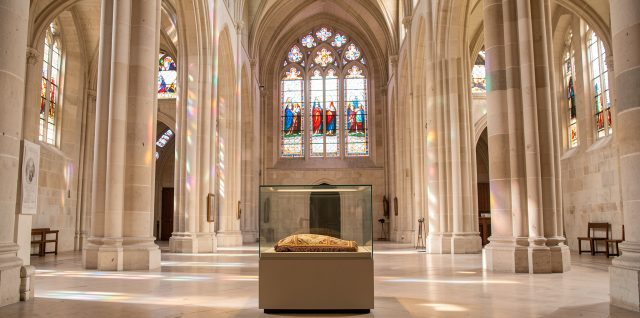 Vue intérieure lumineuse d’une basilique historique avec des vitraux colorés et une relique textile présentée sobrement sous verre protecteur.