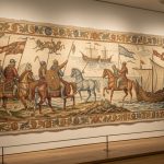 Comment décrypter la Tapisserie de Bayeux et le récit de 1066 ? Tapisserie médiévale brodée sur lin, déployée dans une galerie de musée éclairée par une lumière douce, avec des cavaliers normands et des chevaux représentés dans des tons ocre, rouille, indigo et vert.